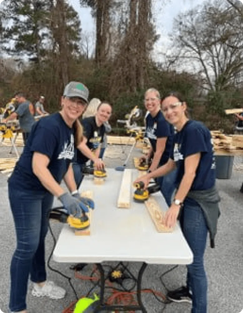 People sanding wood on tables outdoors, surrounded by trees and woodworking tools.