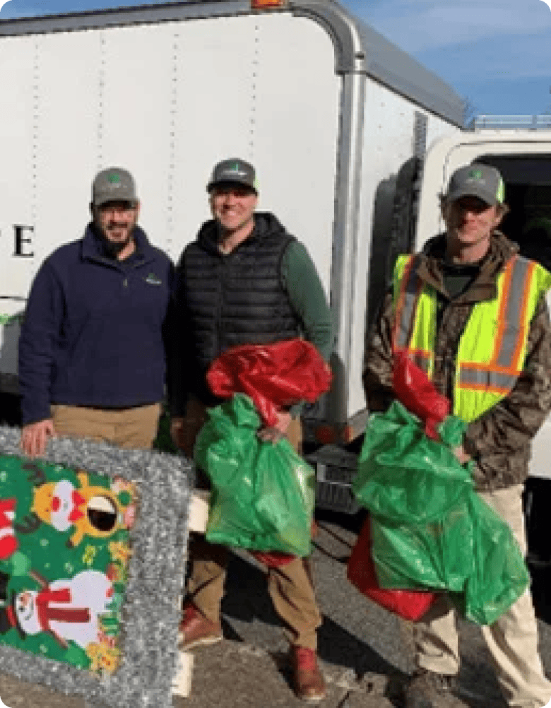 Three men smile holding red and green bags, standing in front of a white truck, with a child obscured by a large gift bag in the foreground.