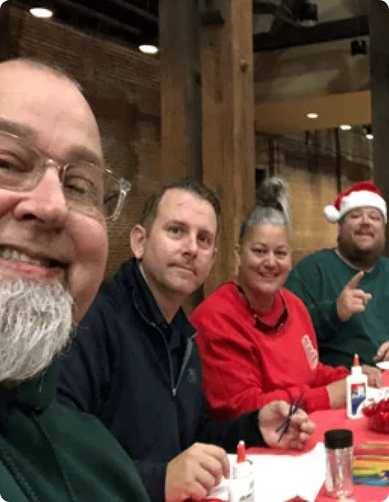 Four adults are seated at a table indoors, smiling for a group photo. Two men wear festive hats, suggesting a holiday gathering.