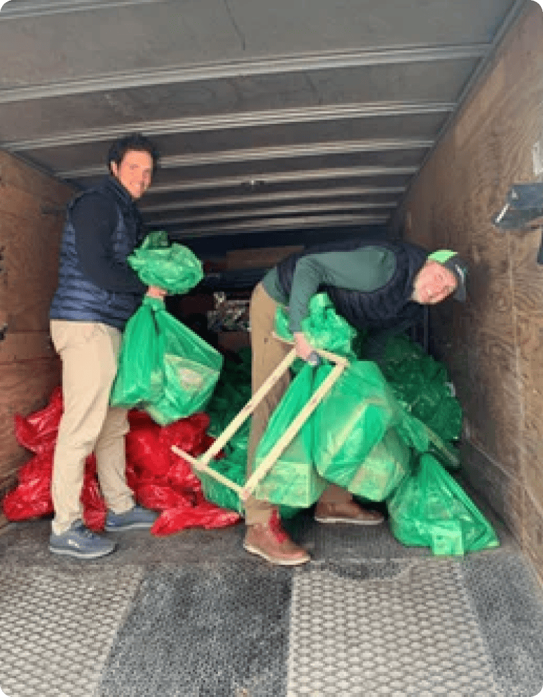 Two men are organizing green bags in a storage space, probably after a clean-up activity, surrounded by a truck's cargo area.