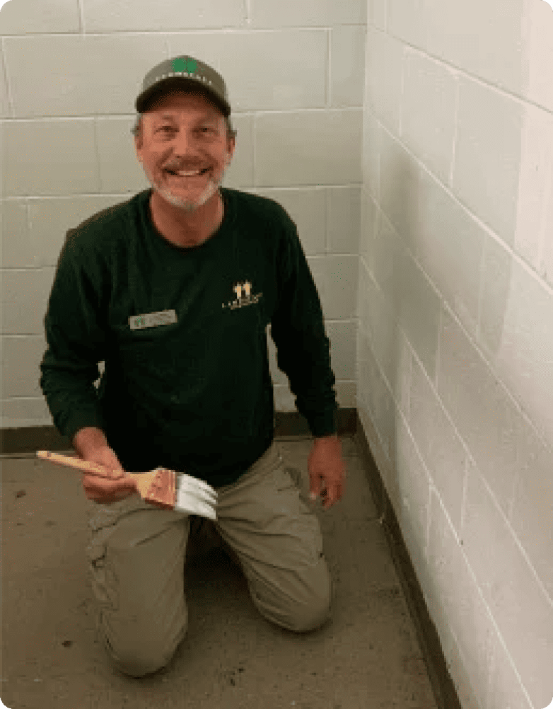 Man kneeling, holding paintbrush, smiling in a room with white brick walls. No text present.