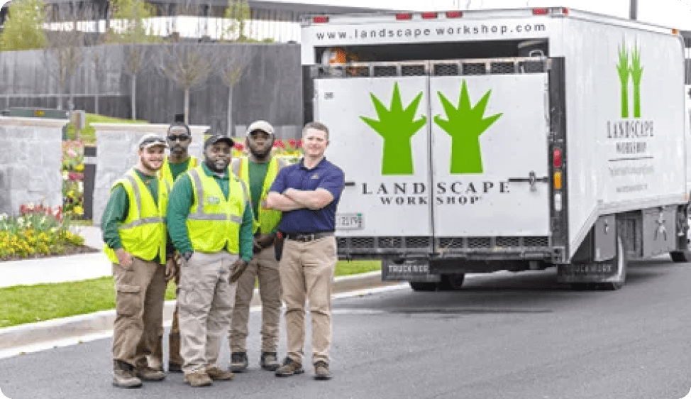 Five men pose together near a white Landscape Workshop truck with green tree graphics. They are likely landscaping professionals standing in a daytime outdoor setting.