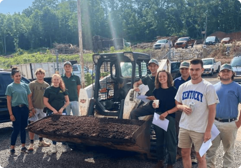 Seven people pose, smiling, with papers and a skid steer loader holding mulch, in an outdoor industrial setting with vehicles and equipment in the background.