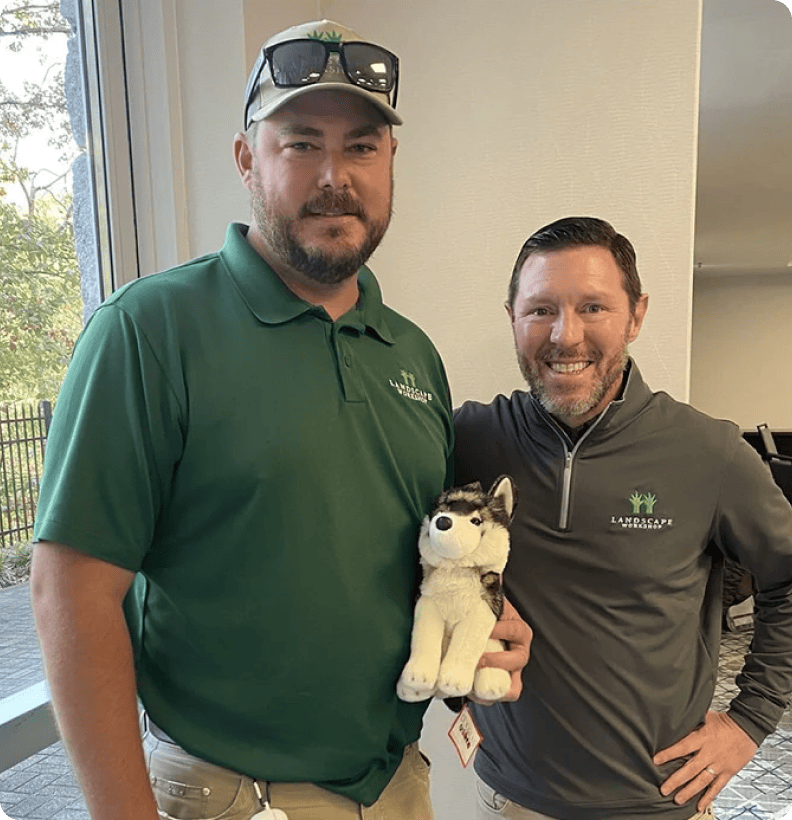 Two smiling men in branded polo shirts pose indoors, one holding a plush husky toy.