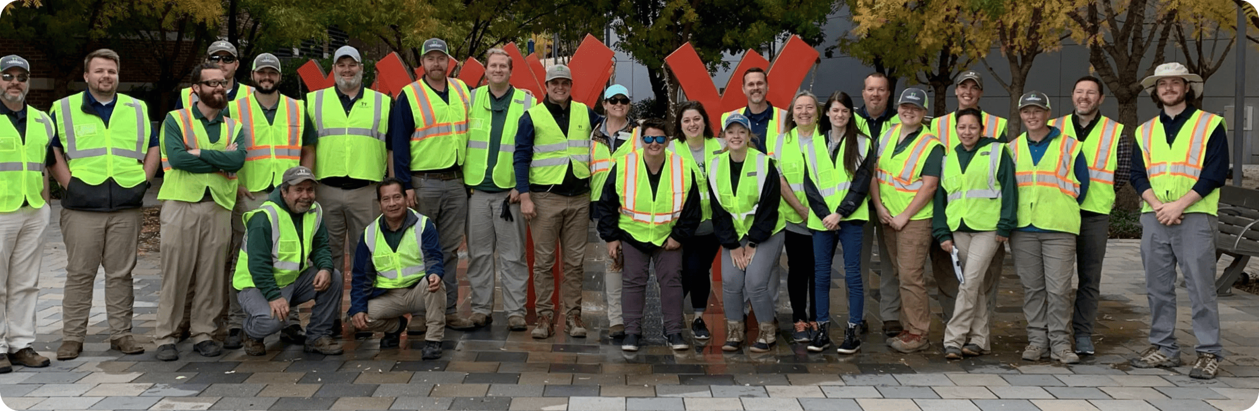 A diverse group of people wearing fluorescent safety vests pose for a photo, standing and squatting in urban outdoor setting with trees and artwork.