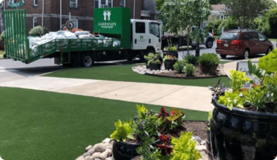 A green landscaping truck is parked on a street, unloading supplies next to a neatly kept garden with various plants and a footpath.