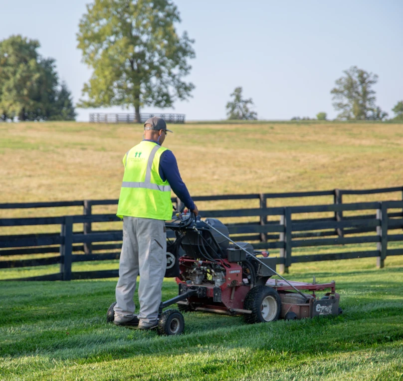 Man on a lawnmower on green grass with black fence and field in the background