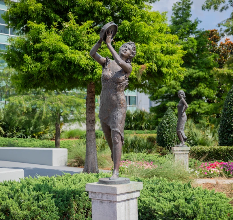 Bronze statue of a woman on a pedestal; trees and bushes in background.