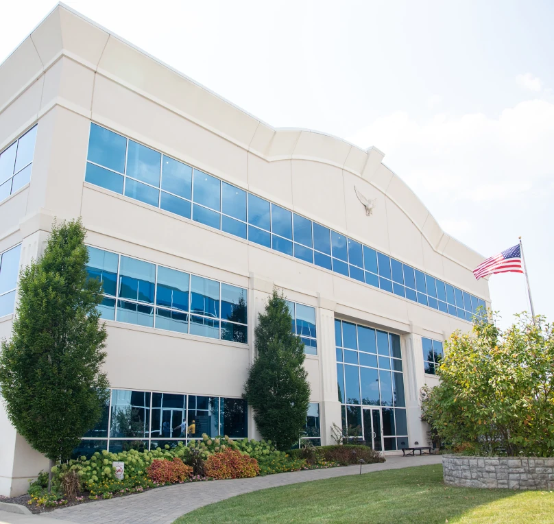 Government building with glass windows, an American flag, paved paths, and shrubs.