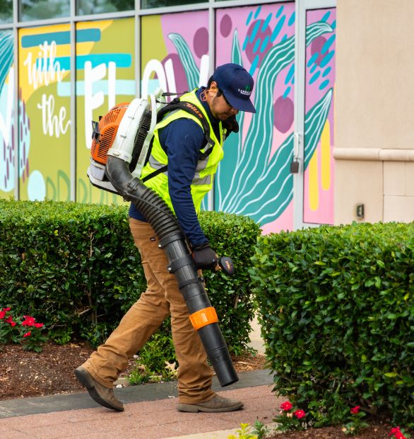 A man in a hi-vis vest carries a large hose leaf blower in front of a business.